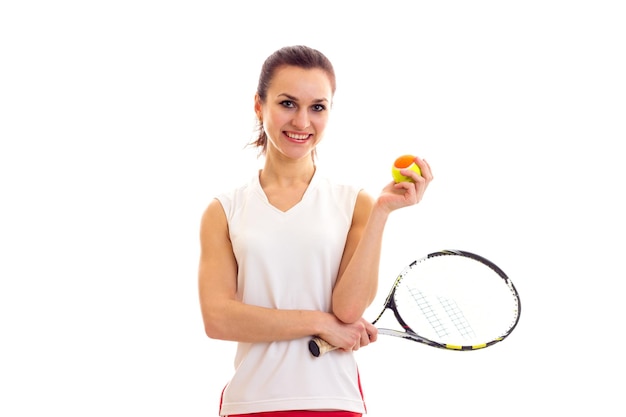 Giovane donna positiva in camicia sportiva bianca e gonna rossa che tiene una racchetta da tennis e una palla gialla