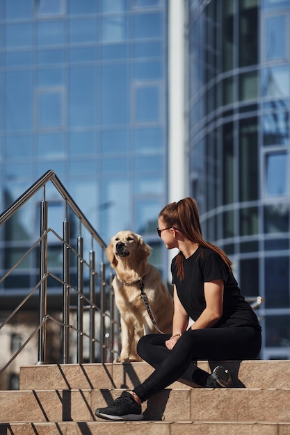 Giovane donna positiva seduta sulle scale con il suo cane quando fa una passeggiata all'aperto vicino all'edificio degli affari