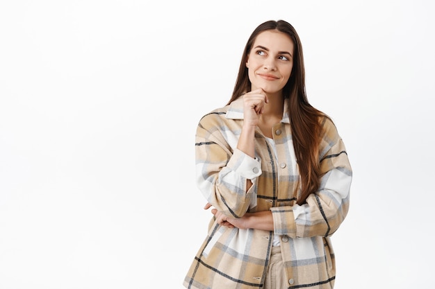 Giovane donna premurosa che guarda da parte e immagina, immagina o pensa a qualcosa di interessante, sorridente pensierosa, in piedi su un muro bianco