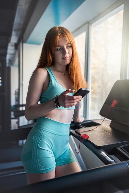 Giovane donna redhair in piedi vicino ai trainer del tapis roulant che utilizzano la cella mobile in palestra Stile di vita di allenamento