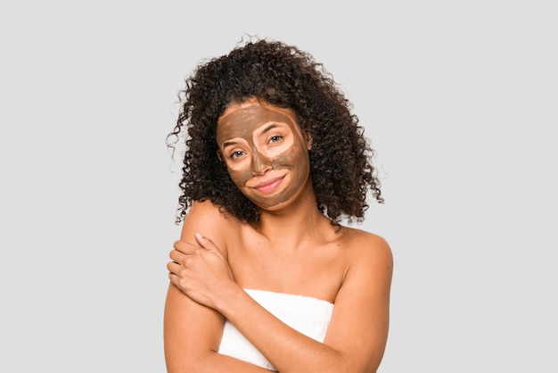 Giovane donna riccia afroamericana che indossa un asciugamano e una maschera facciale isolata