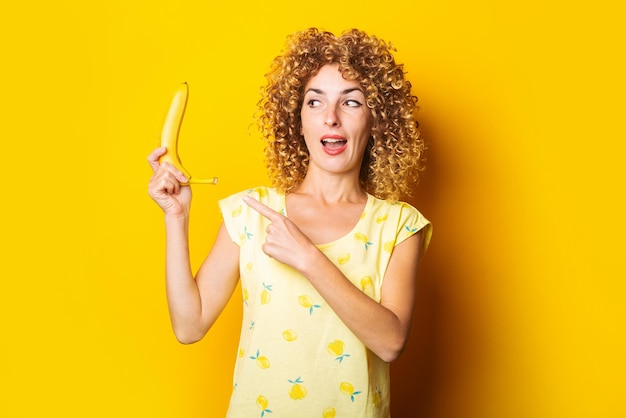 Giovane donna riccia che tiene una banana che punta il dito su uno sfondo giallo