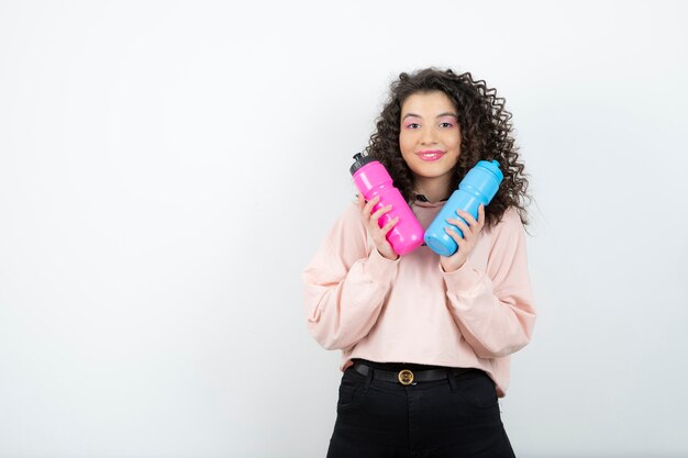 Giovane donna riccia in maglione che tiene bottiglie d'acqua rosa e blu.