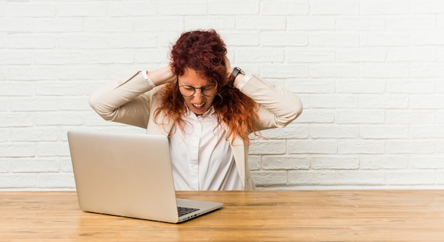 Giovane donna riccia rossa che lavora con il suo computer portatile che copre le orecchie con le mani cercando di non sentire un suono troppo forte.