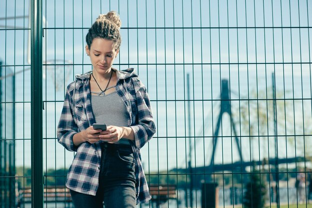 Giovane donna rilassata che indossa abiti casual e sorride leggermente mentre guarda lo schermo del suo smartphone moderno