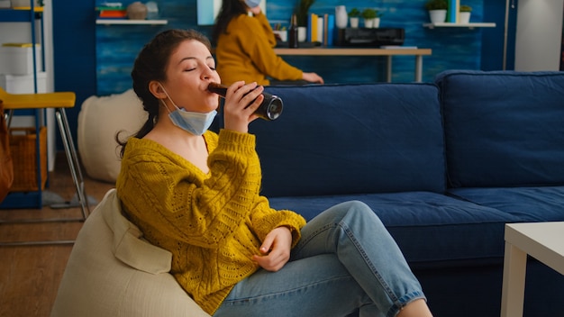 Giovane donna rilassata che si toglie la maschera facciale che guarda la telecamera bevendo birra seduta sul divano mentre passa il tempo con gli amici durante la pandemia globale con il coronavirus. Gruppo di giovani che si divertono.