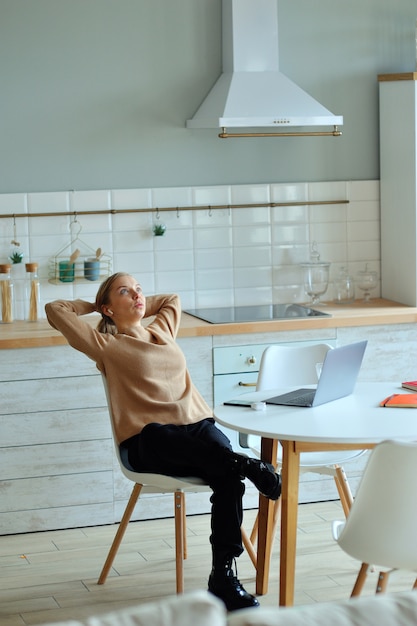 Giovane donna rilassata felice che si siede nella sua cucina con un computer portatile