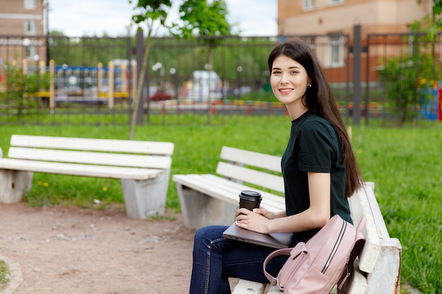 Giovane donna rilassata in una maglietta verde e con uno zaino che si siede su un banco di legno mentre bevendo caffè e navigando sul suo computer portatile