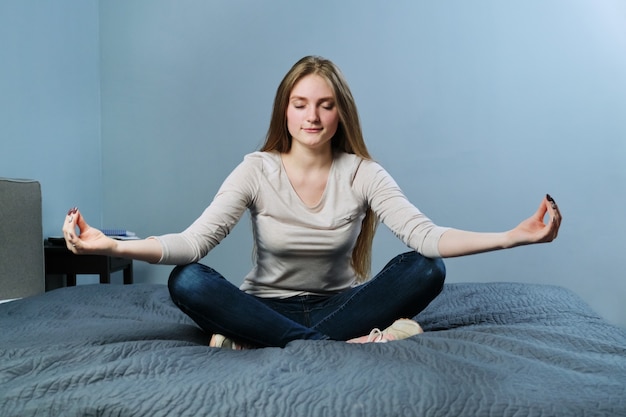 Giovane donna rilassata seduta a casa a letto nella posizione del loto, meditando ragazza che pratica yoga