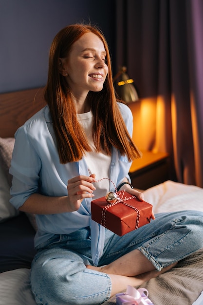 Giovane donna rossa allegra che apre il contenitore di regalo con il presente con gli occhi chiusi nell'accogliente soggiorno vicino al computer portatile. Donna sognante che ha una chat video online seduta a letto in una camera da letto leggera.