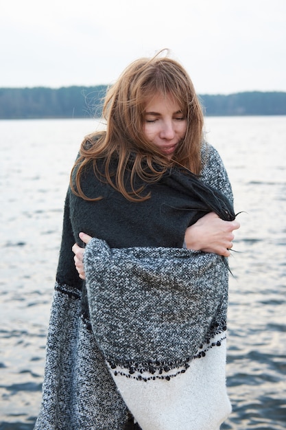 Giovane donna rossa avvolta in una coperta plaid sorridente, in piedi vicino al lago. Brutto tempo nuvoloso ventoso freddo autunnale.