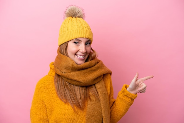 Giovane donna rossa che indossa una giacca invernale isolata su sfondo rosa che punta il dito di lato