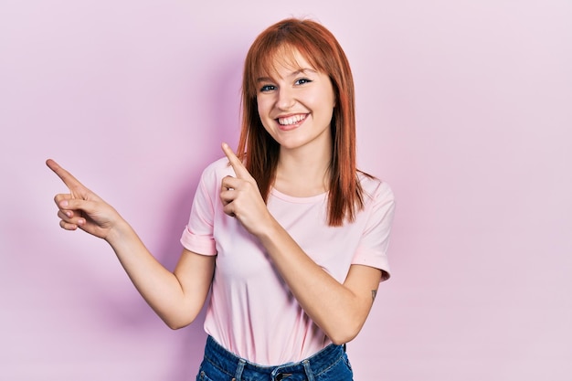 Giovane donna rossa che indossa una maglietta rosa casual sorridendo e guardando la telecamera che punta con due mani e dita di lato