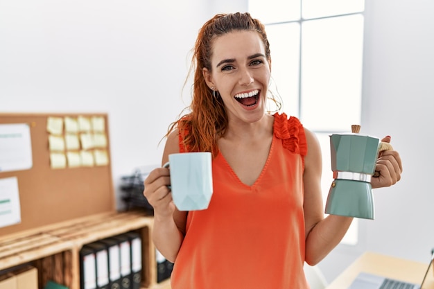 Giovane donna rossa che tiene la caffettiera francese in ufficio sorridendo e ridendo ad alta voce perché divertente scherzo pazzo.