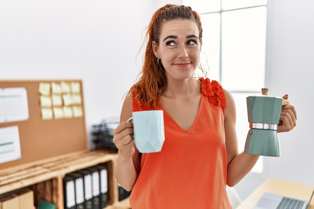 Giovane donna rossa che tiene la caffettiera francese in ufficio sorridendo guardando di lato e fissando lontano pensando