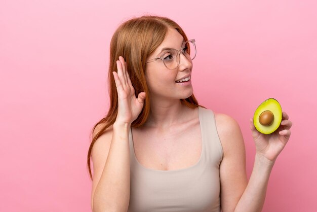 Giovane donna rossa che tiene un avocado isolato su sfondo rosa ascoltando qualcosa mettendo la mano sull'orecchio