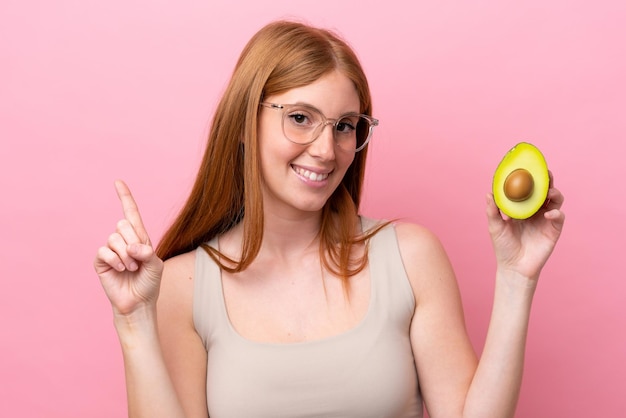 Giovane donna rossa che tiene un avocado isolato su sfondo rosa che mostra e alza un dito in segno di meglio