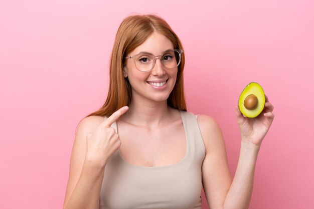 Giovane donna rossa che tiene un avocado isolato su sfondo rosa dando un pollice in alto gesto