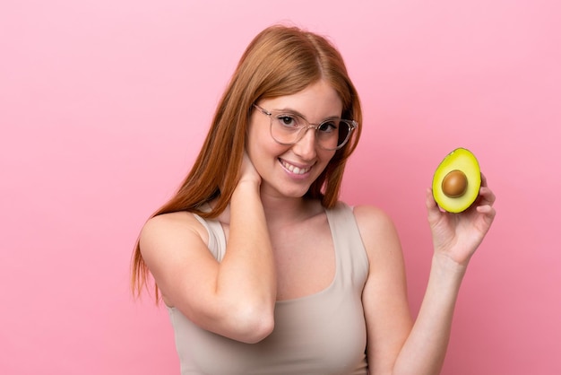 Giovane donna rossa che tiene un avocado isolato su sfondo rosa ridendo