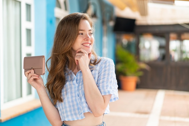 Giovane donna rossa che tiene un portafoglio all'aperto pensando a un'idea e guardando un lato