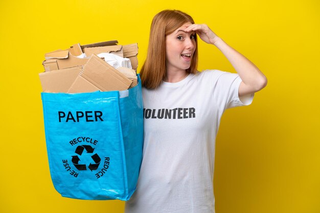 Giovane donna rossa che tiene un sacchetto di riciclaggio pieno di carta da riciclare isolato su sfondo giallo facendo un gesto a sorpresa mentre guarda di lato
