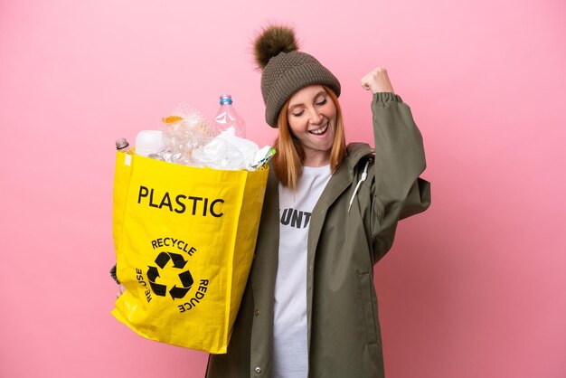 Giovane donna rossa che tiene un sacchetto pieno di bottiglie di plastica da riciclare isolato su sfondo rosa che celebra una vittoria