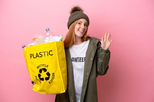 Giovane donna rossa che tiene un sacchetto pieno di bottiglie di plastica da riciclare isolato su sfondo rosa salutando con la mano con espressione felice