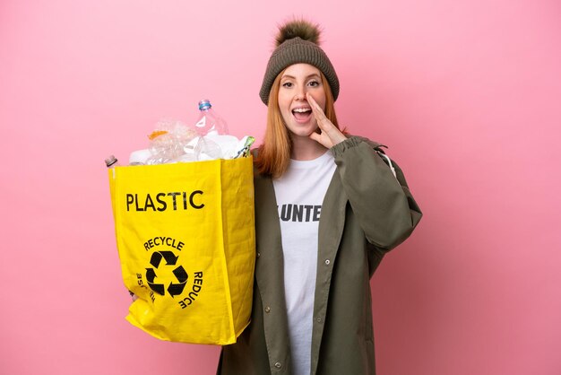 Giovane donna rossa che tiene una borsa piena di bottiglie di plastica da riciclare isolata su sfondo rosa che grida con la bocca spalancata