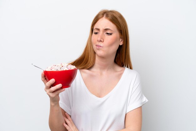 Giovane donna rossa che tiene una ciotola di cereali isolata su sfondo bianco con espressione triste
