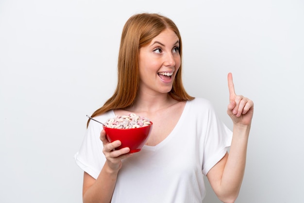 Giovane donna rossa che tiene una ciotola di cereali isolata su sfondo bianco con l'intenzione di realizzare la soluzione sollevando un dito
