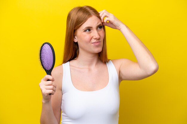 Giovane donna rossa che tiene una spazzola per capelli isolata su sfondo bianco con dubbi e con un'espressione facciale confusa