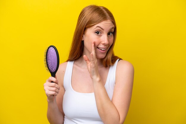 Giovane donna rossa che tiene una spazzola per capelli isolata su sfondo bianco sussurrando qualcosa