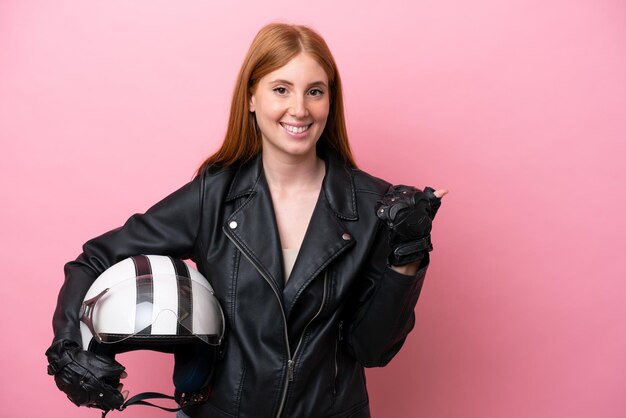 Giovane donna rossa con un casco da motociclista isolato su sfondo rosa che punta di lato per presentare un prodotto