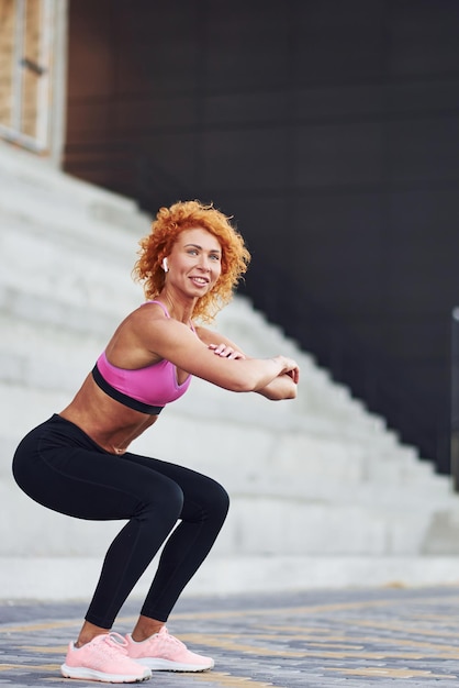 Giovane donna rossa europea in abiti sportivi che fanno squat all'aperto