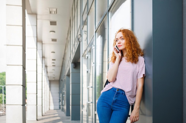 Giovane donna rossa felice che chiama sul telefono cellulare sulla strada per le classi. Istruzione, tecnologia, comunicazione e concetto di connessione. Copia spazio sul muro