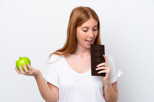 Giovane donna rossa isolata su sfondo bianco che prende una tavoletta di cioccolato in una mano e una mela nell'altra