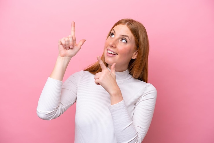Foto giovane donna rossa isolata su sfondo rosa che punta con il dito indice una grande idea
