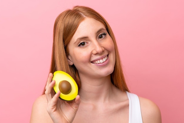 Giovane donna rossa isolata su sfondo rosa che tiene un avocado mentre sorride Primo piano ritratto