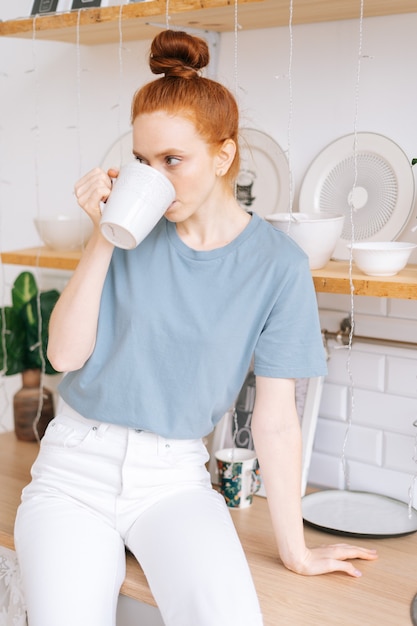 Giovane donna rossa rilassata che tiene una tazza di caffè caldo mentre sta in piedi nella stanza della cucina