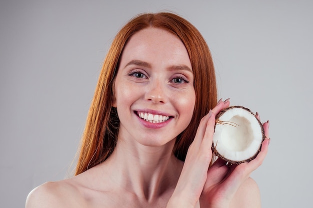 Giovane donna rossa rossa con pelle ideale e capelli rossi perfetti che tiene in mano una noce di cocco in uno sfondo bianco dello studio.