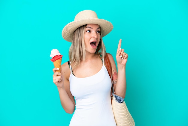 Giovane donna rumena che tiene gelato e borsa da spiaggia isolata su sfondo blu con l'intenzione di realizzare la soluzione sollevando un dito