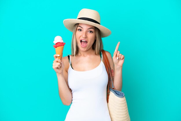 Giovane donna rumena che tiene il gelato e la borsa da spiaggia isolata su sfondo blu che punta verso l'alto una grande idea