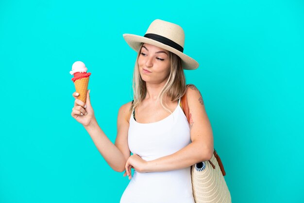 Giovane donna rumena che tiene il gelato e la borsa da spiaggia isolati su sfondo blu con espressione triste