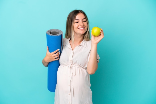 Giovane donna rumena isolata su sfondo blu incinta che tiene una mela e va a lezioni di yoga