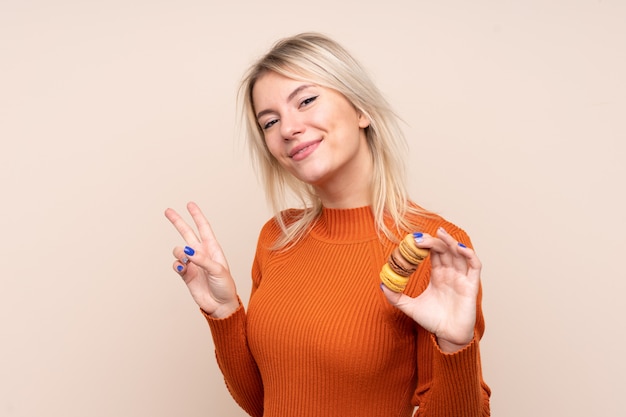 Giovane donna russa bionda sopra la parete isolata che tiene i macarons francesi variopinti e che mostra il segno di vittoria