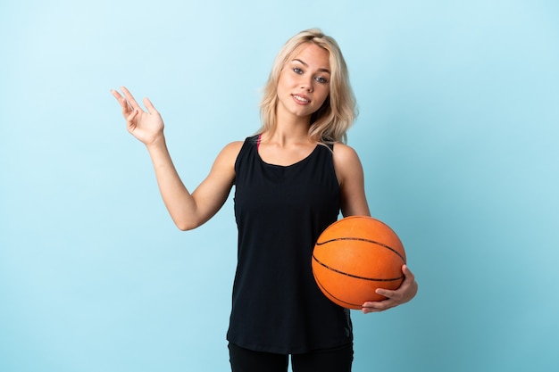 Giovane donna russa che gioca a basket isolato su blu che estende le mani a lato per invitare a venire