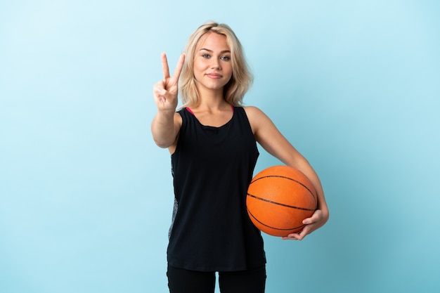 Giovane donna russa che gioca a basket isolato sulla parete blu sorridendo e mostrando il segno di vittoria