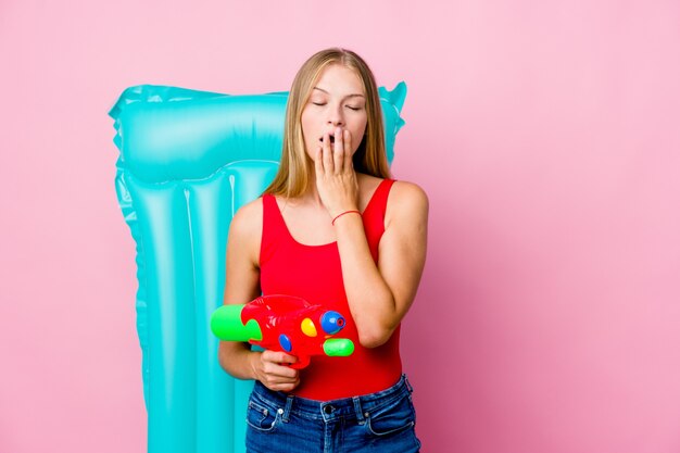 Giovane donna russa che gioca con una pistola ad acqua con un materasso ad aria che sbadiglia mostrando un gesto stanco che copre la bocca con la mano.