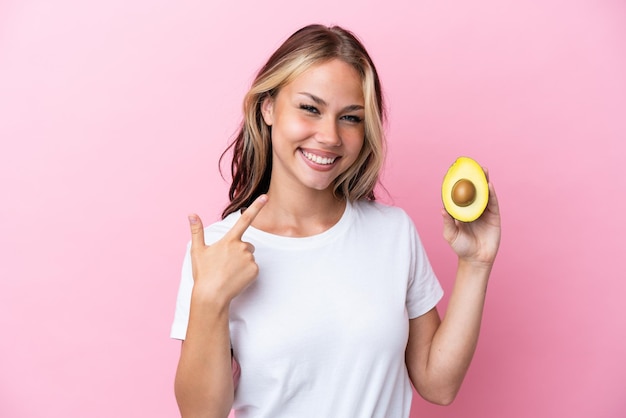 Giovane donna russa che tiene avocado isolato su sfondo rosa dando un pollice in alto gesto