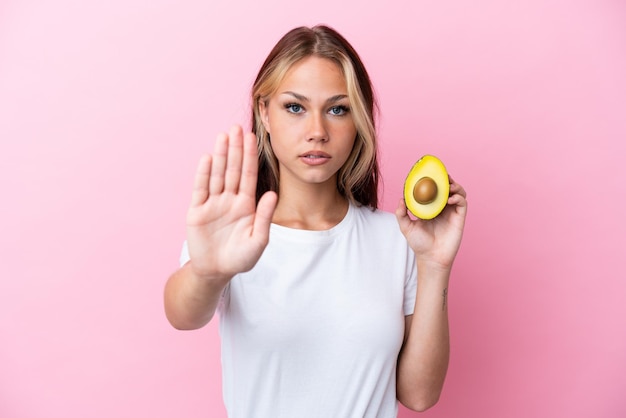 Giovane donna russa che tiene avocado isolato su sfondo rosa facendo un gesto di arresto